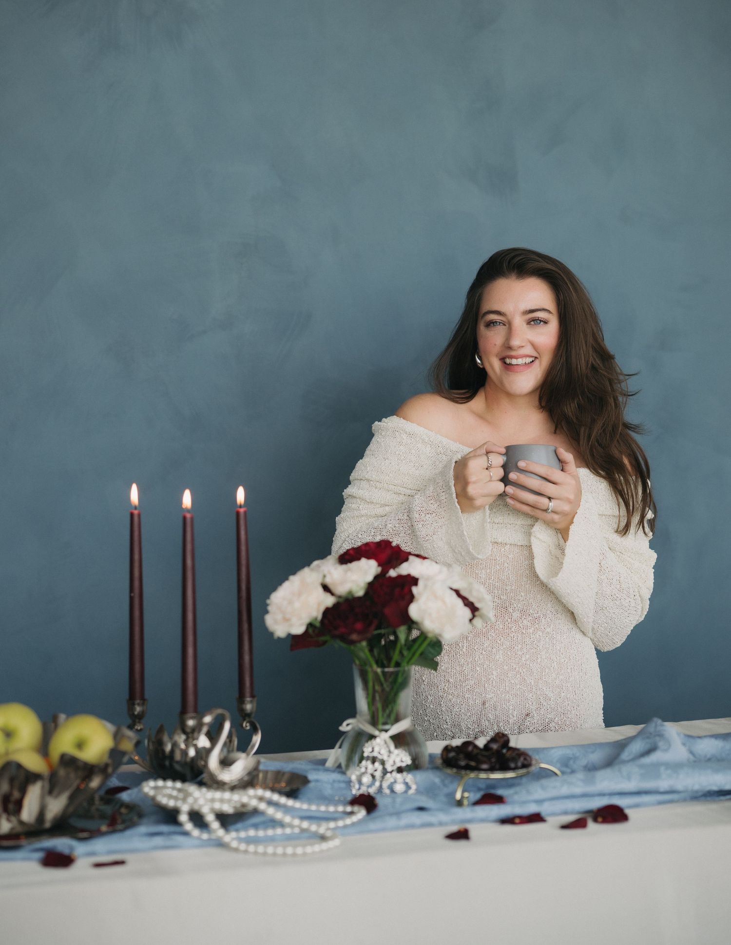A cozy winter scene with lit candles, flowers, and someone in a white sweater relaxing at a decorated table setting.