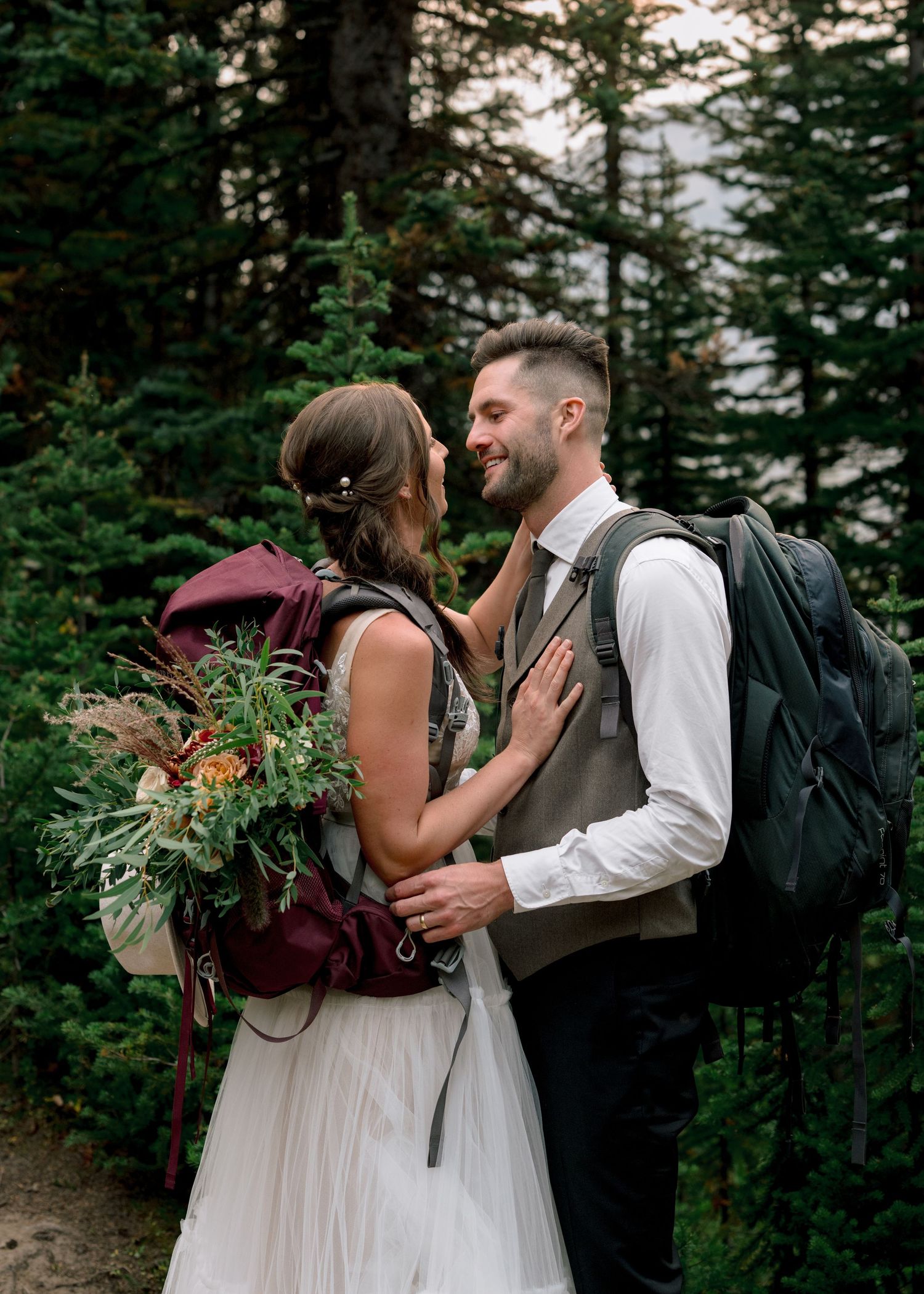 A romantic mountain elopement scene with two backpack-wearing individuals embracing among evergreen trees at sunset.