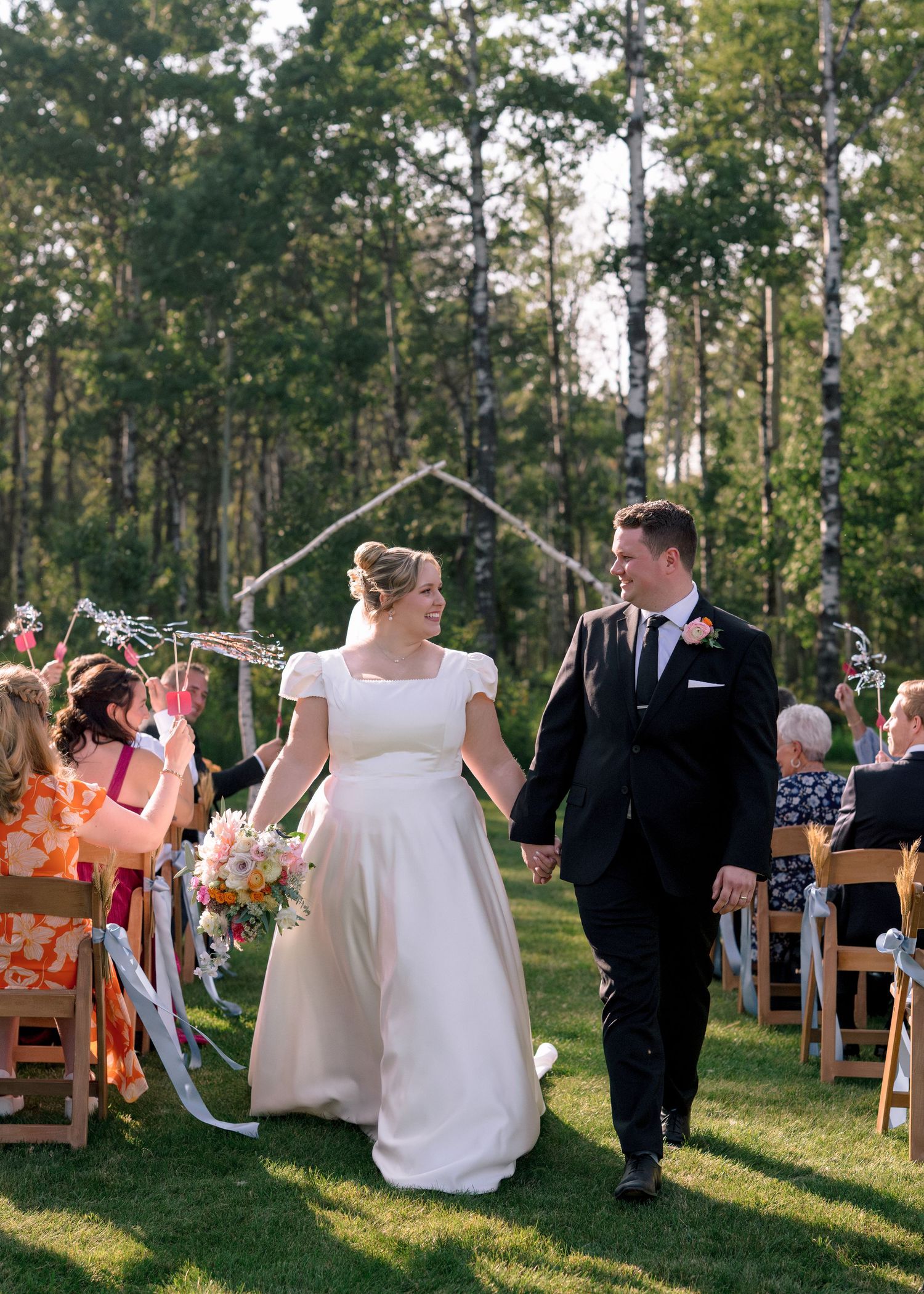 Newlyweds walk down the aisle together at an outdoor woodland wedding ceremony as guests look on from wooden chairs.