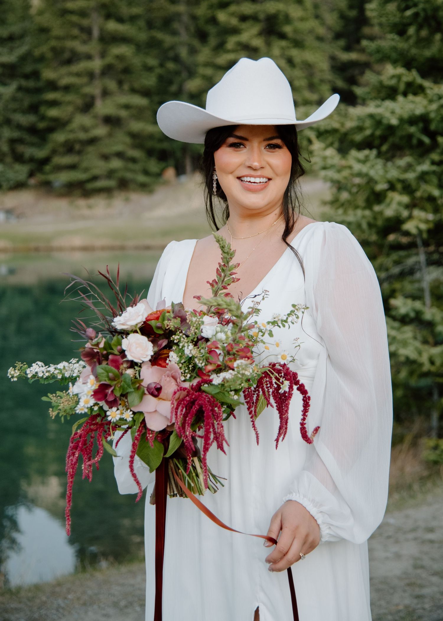 A bride in a flowing white dress and cowboy hat holds a vibrant burgundy and pink bouquet. Banff and Calgary Wedding Photographer