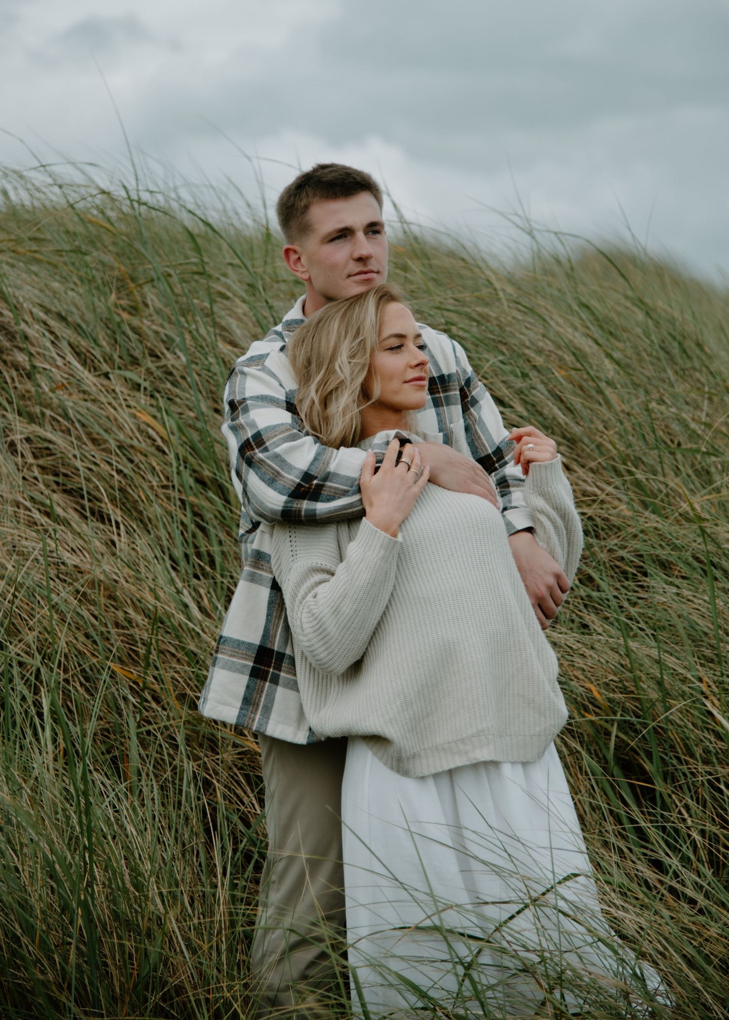 A tender embrace between a couple standing in wind-swept coastal grass on Oregon Coast. Calgary Lifestyle and Engagement Photographer.