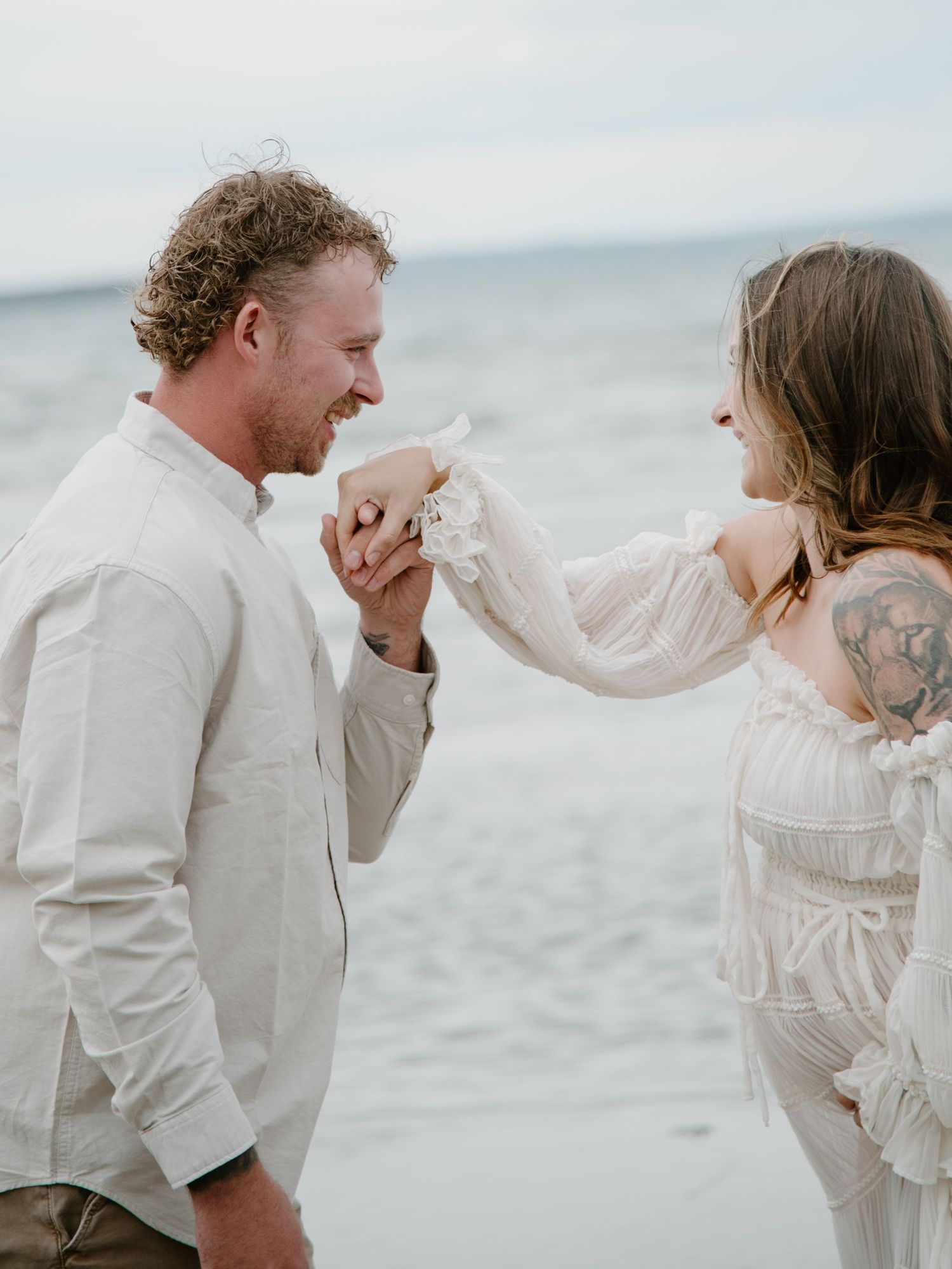 A joyful moment between a couple in matching light attire on a scenic beach. Calgary Alberta Maternity Photographer.