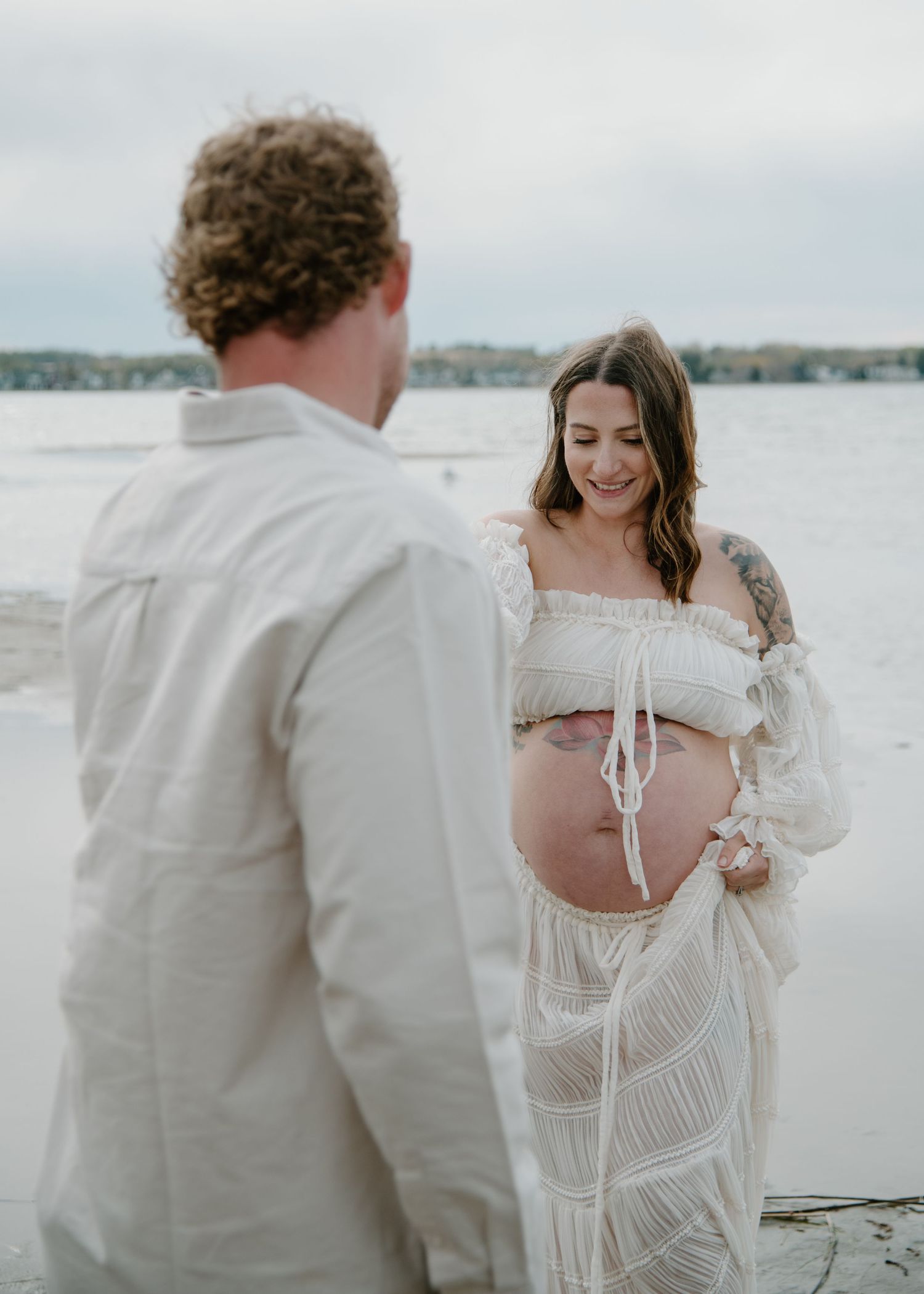 Intimate waterside maternity photo with couple wearing white against a serene coastal backdrop. Calgary Alberta Maternity Photographer.