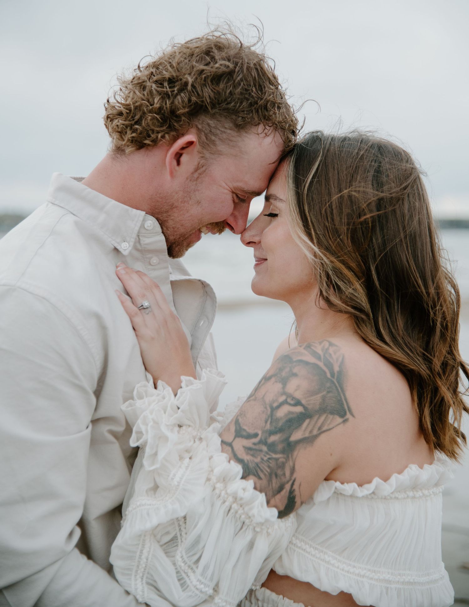 A romantic couple in white outfits shares an intimate moment against a cloudy beach backdrop. Calgary Alberta Maternity Photographer.