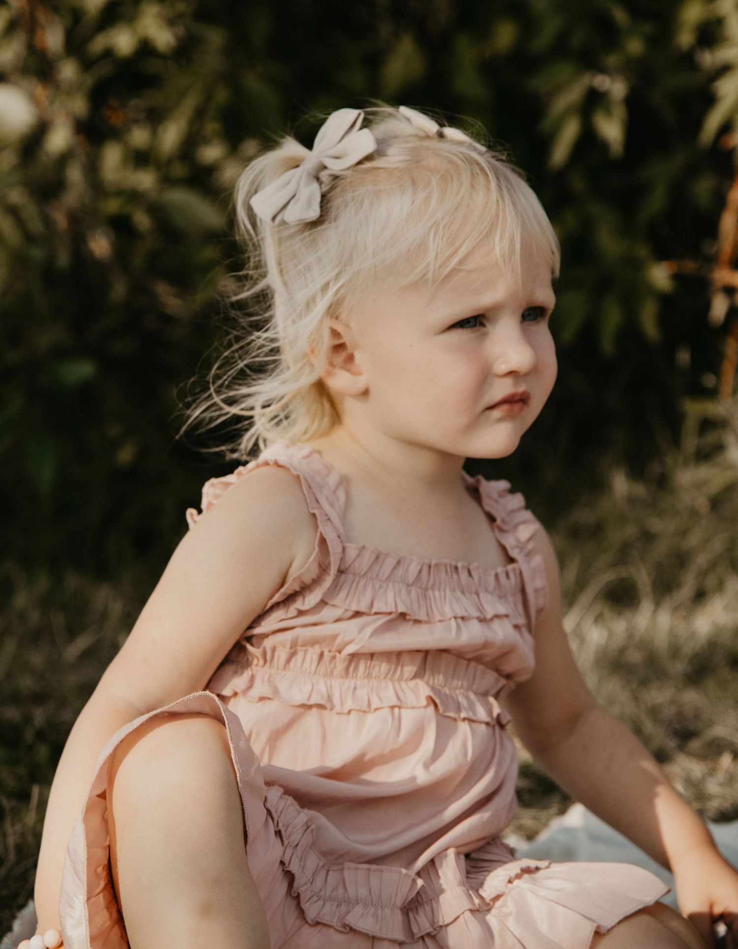A young child in a pink ruffled dress with a bow in their hair sits outdoors. Calgary Alberta Family Photographer.