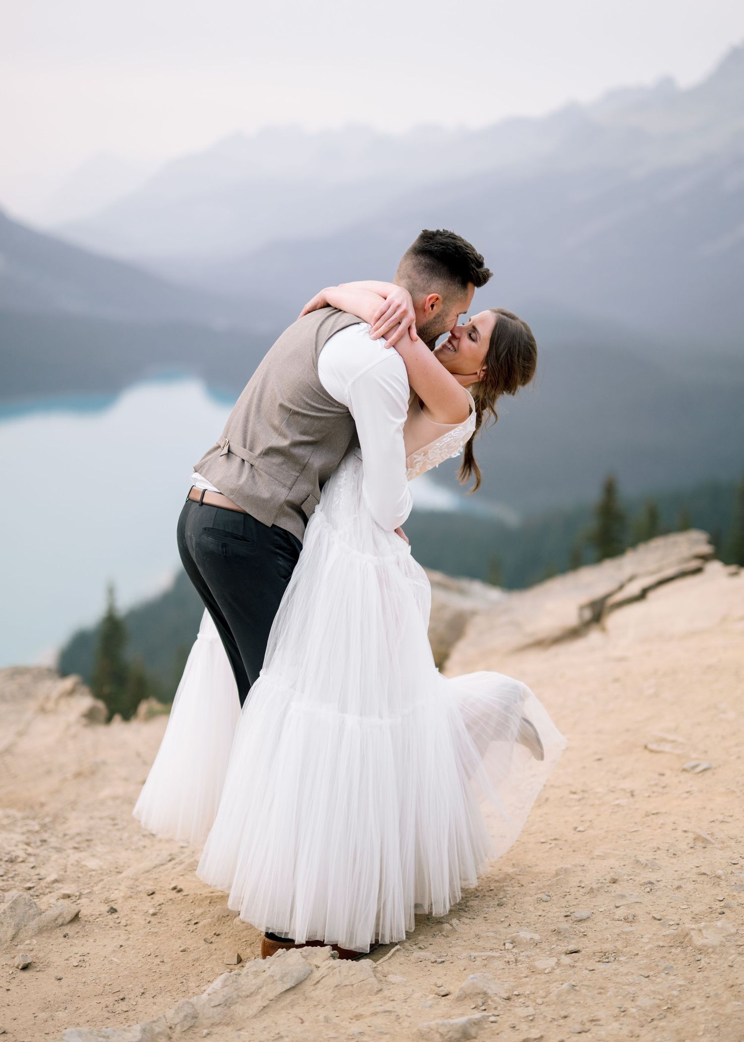 A romantic wedding couple embraces on a mountain cliff with misty peaks and a lake in the background.