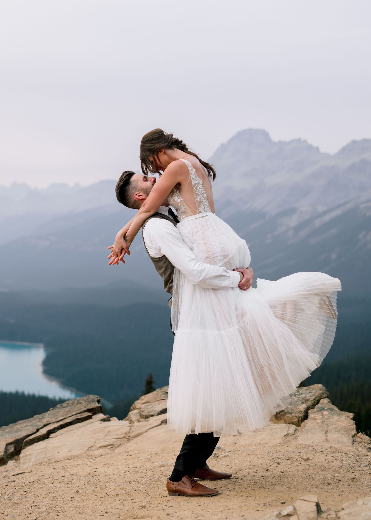 A figure in a white dress poses dramatically on a rocky mountain ledge overlooking a scenic lake and mountain landscape.