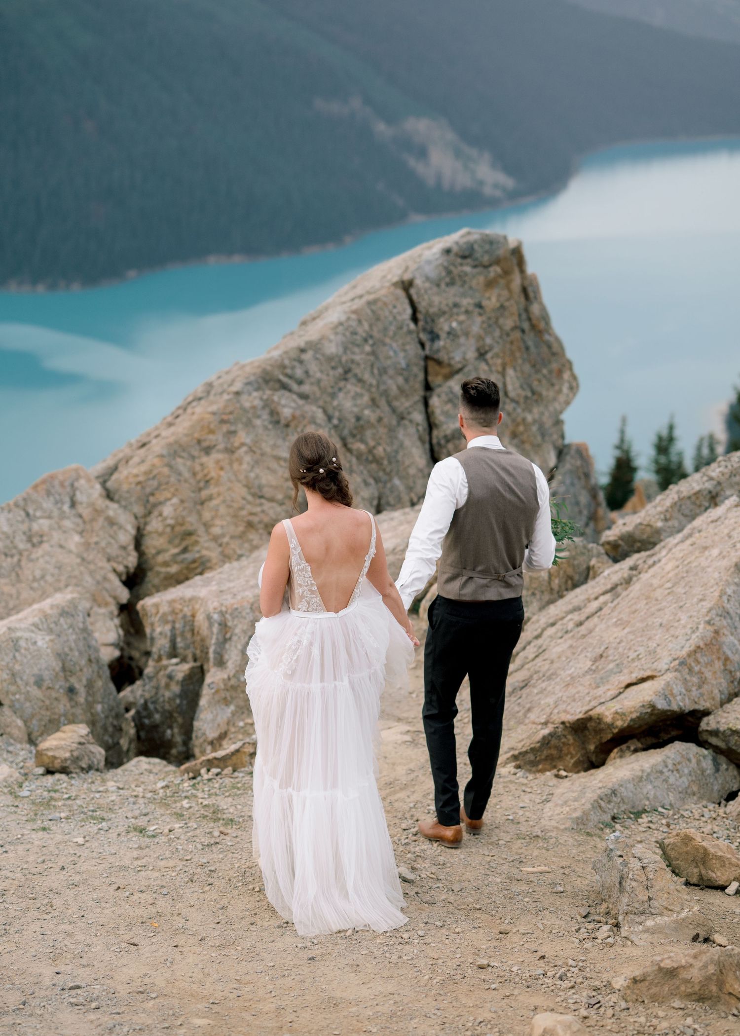 Couple in formal attire walks along a rocky mountain ridge overlooking a turquoise alpine lake on their wedding day.