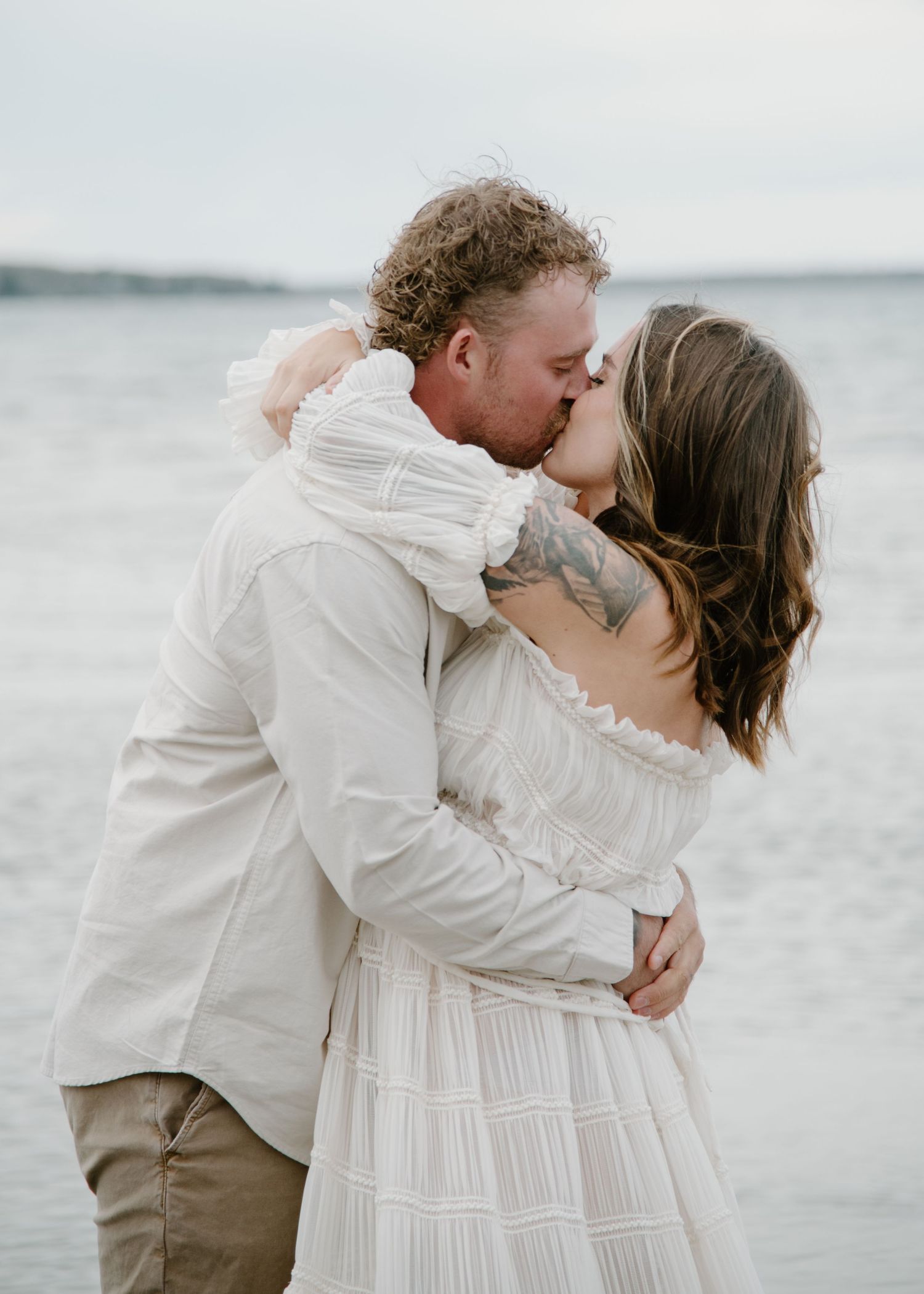 A passionate embrace between a couple wearing white and beige on a misty beach. Calgary Alberta Maternity Photographer.