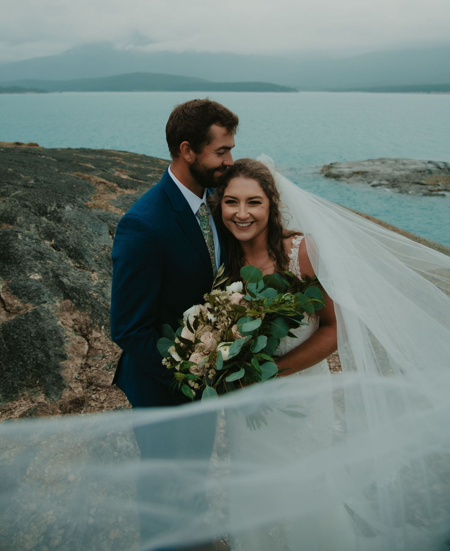 A wedding couple embraces on rocky terrain with a blue lake and mountains in the background. Calgary Wedding Photographer
