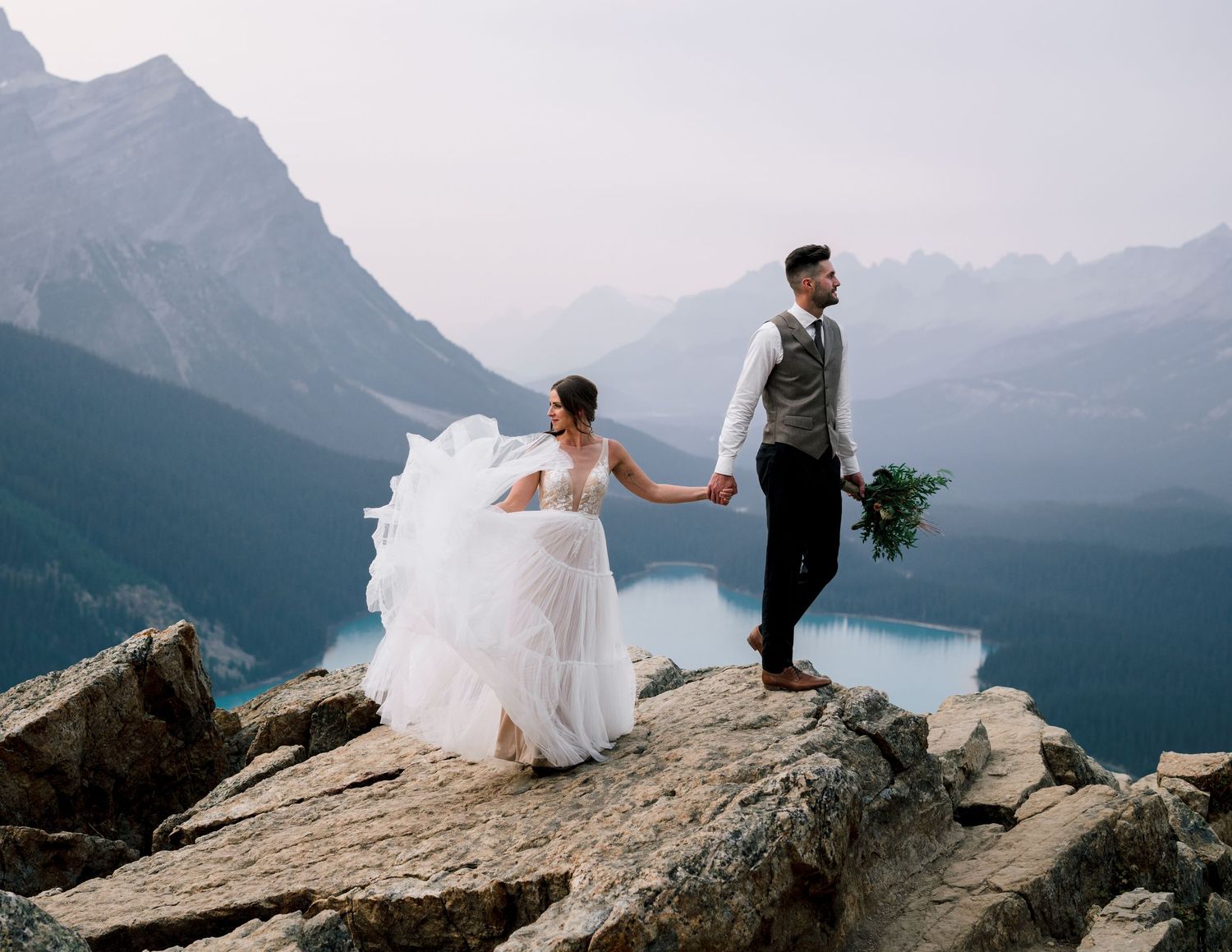 A bride and groom hold hands while standing on a mountain cliff edge with a turquoise lake in the distance.