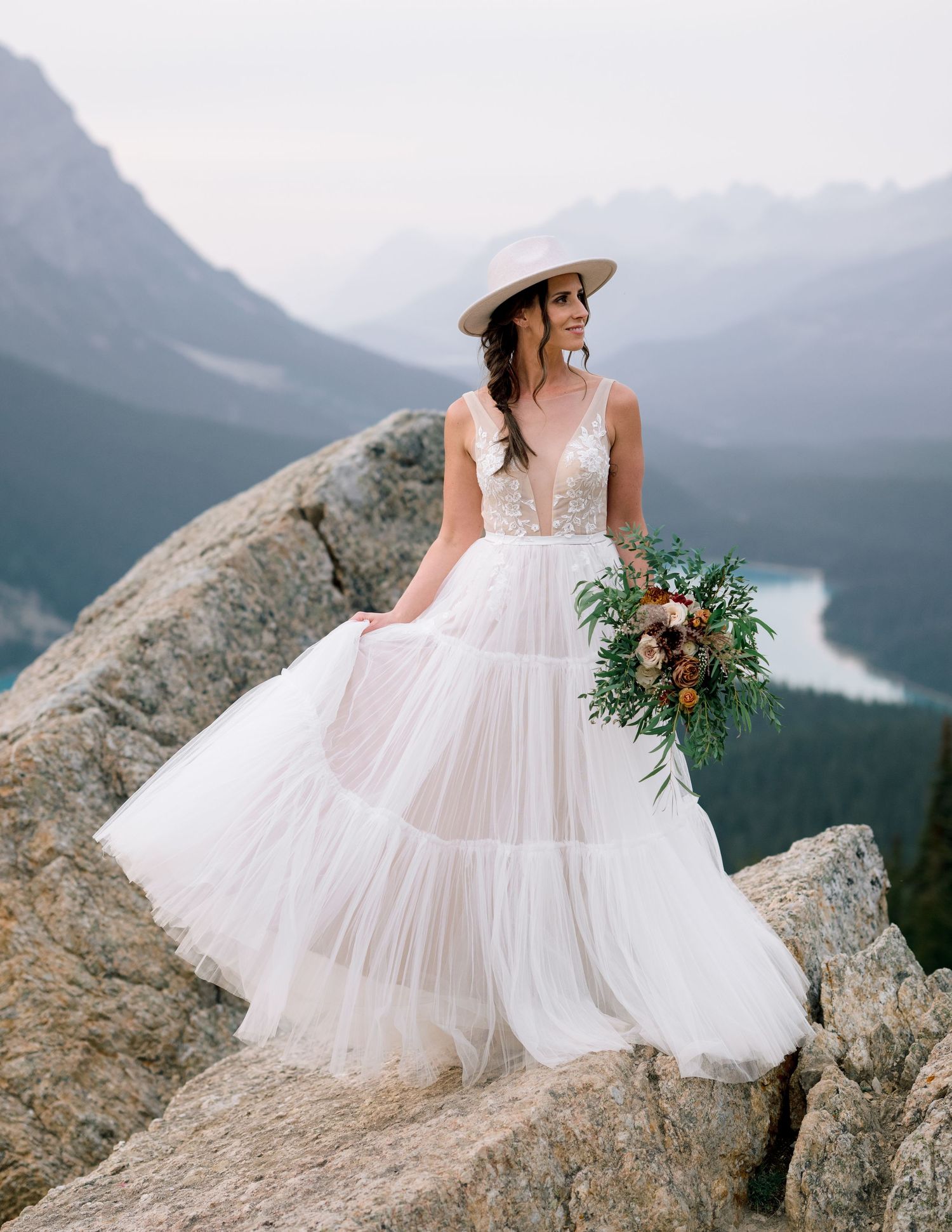 A bride in a flowing white dress and sun hat poses on mountain rocks with distant peaks and valleys behind her.