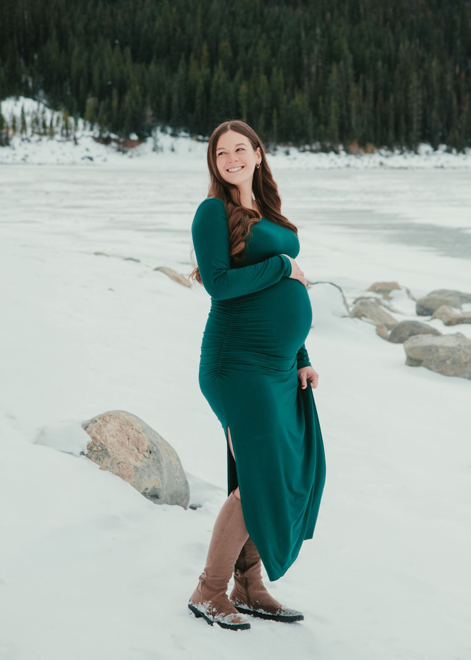 A woman in an emerald green dress poses in front of a snowy mountain landscape. Calgary Alberta Maternity Photographer.