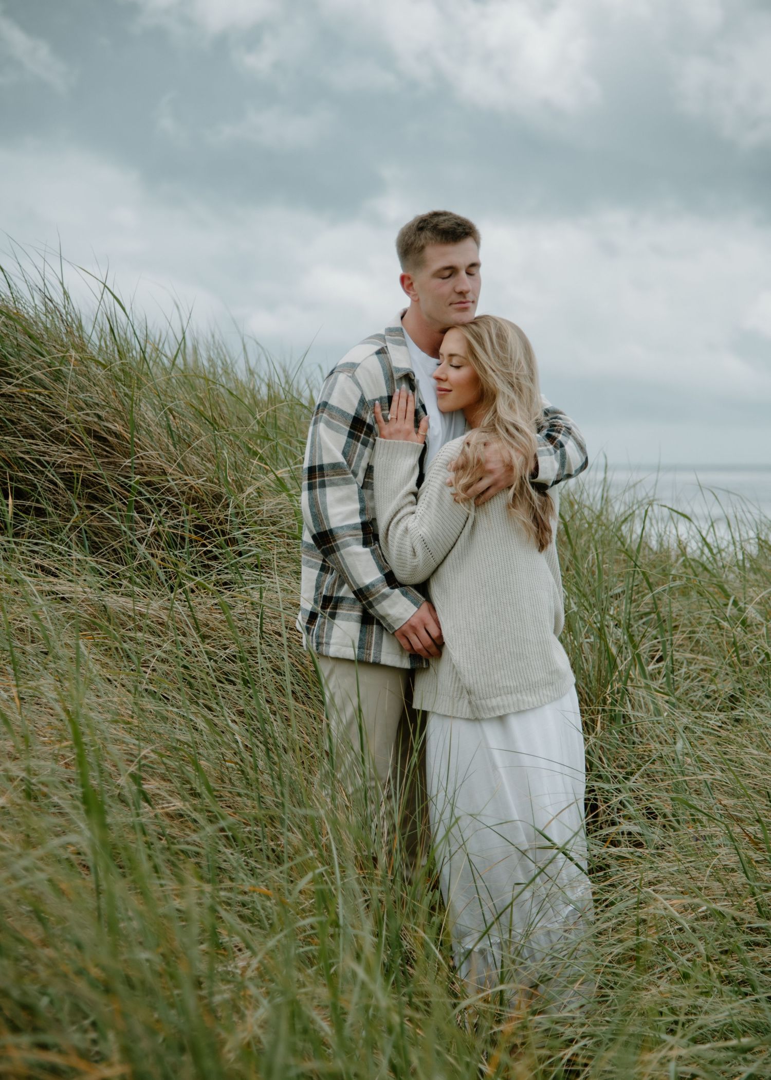 Romantic photoshoot of couple among wild coastal grasses on overcast day on Oregon Coast. Calgary Lifestyle and Engagement Photographer.