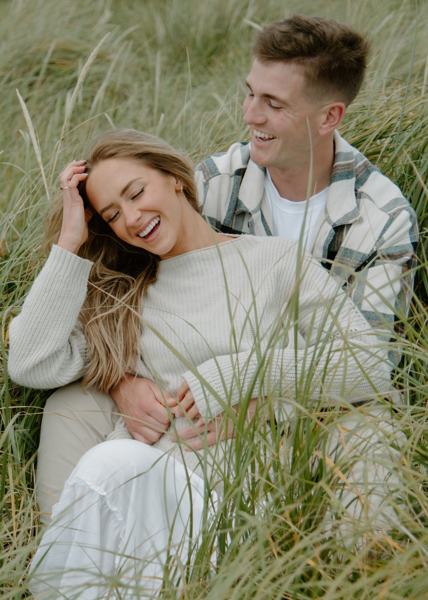 Joyful couple sharing laughter while embracing in tall grass on Oregon Coast. Calgary Lifestyle and Engagement Photographer.