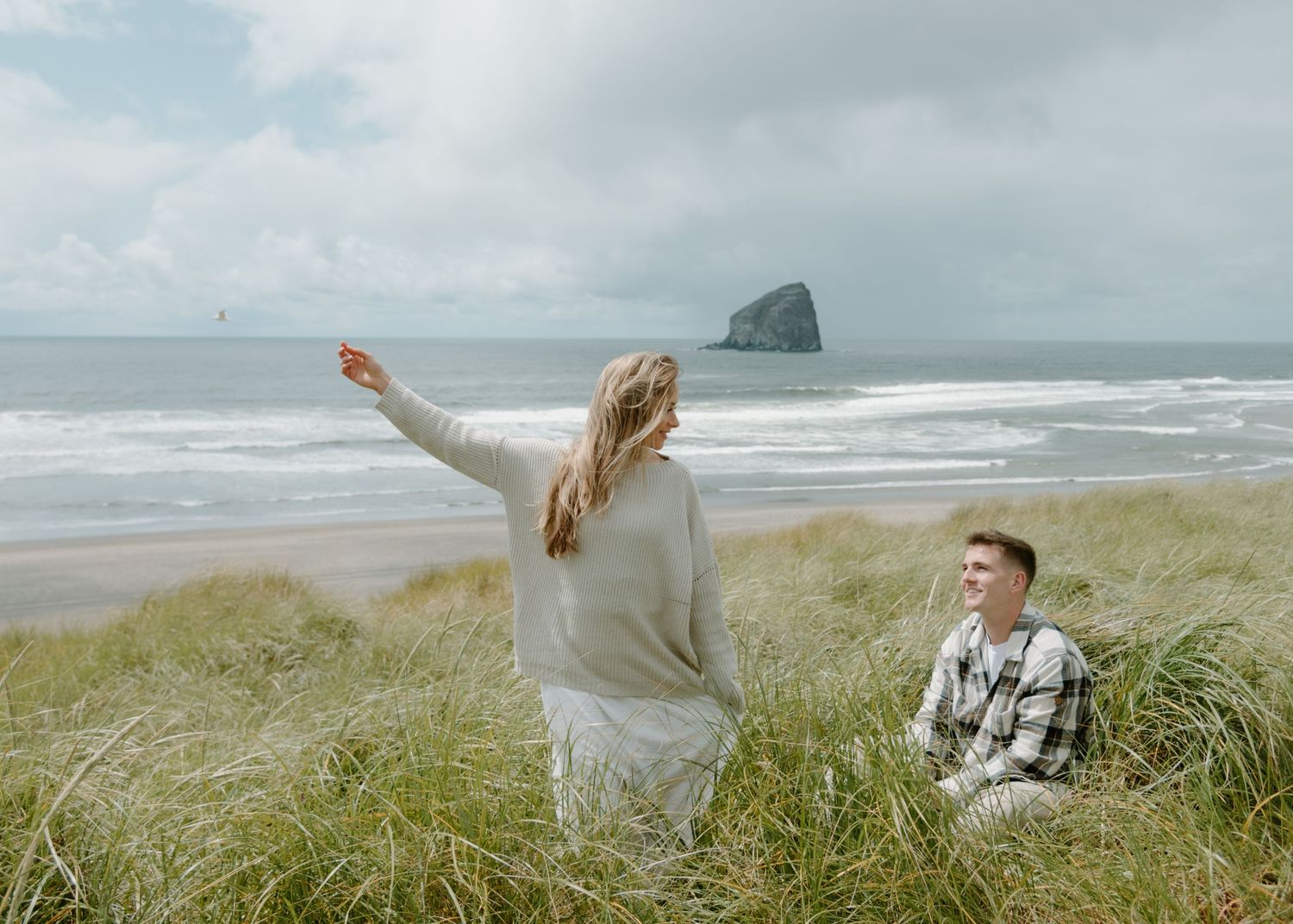 Woman stands with outstretched arms on beach while companion sits in dune grass on Oregon Coast. Calgary Lifestyle and Engagement Photograph