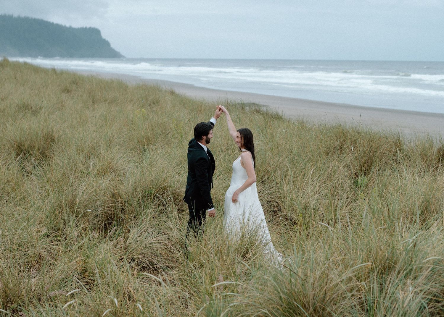 A couple stands amid tall beach grass on a misty Pacific Northwest coastline during a wedding photoshoot.