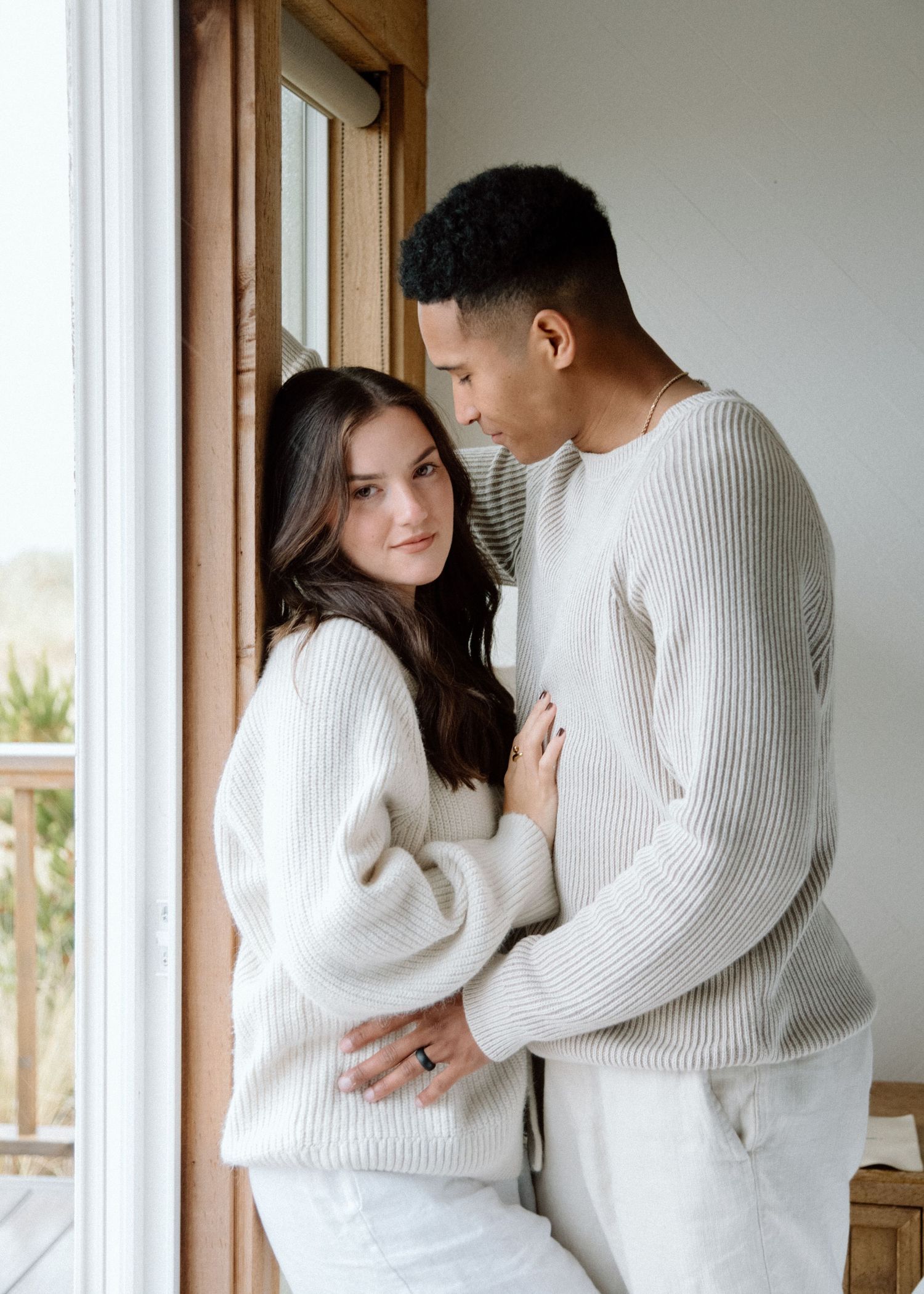Romantic couple wearing matching white sweaters embracing near a bright window.