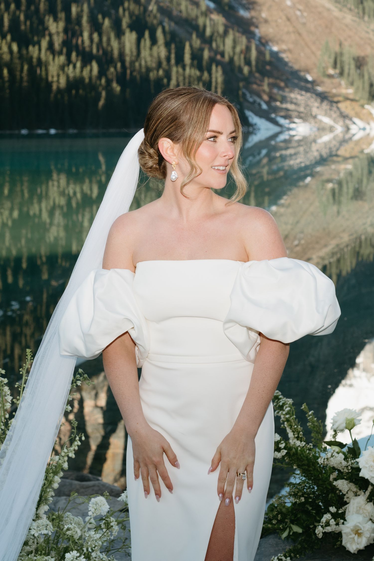 Bride in an elegant off-shoulder white dress poses against a scenic mountain lake backdrop in nature.