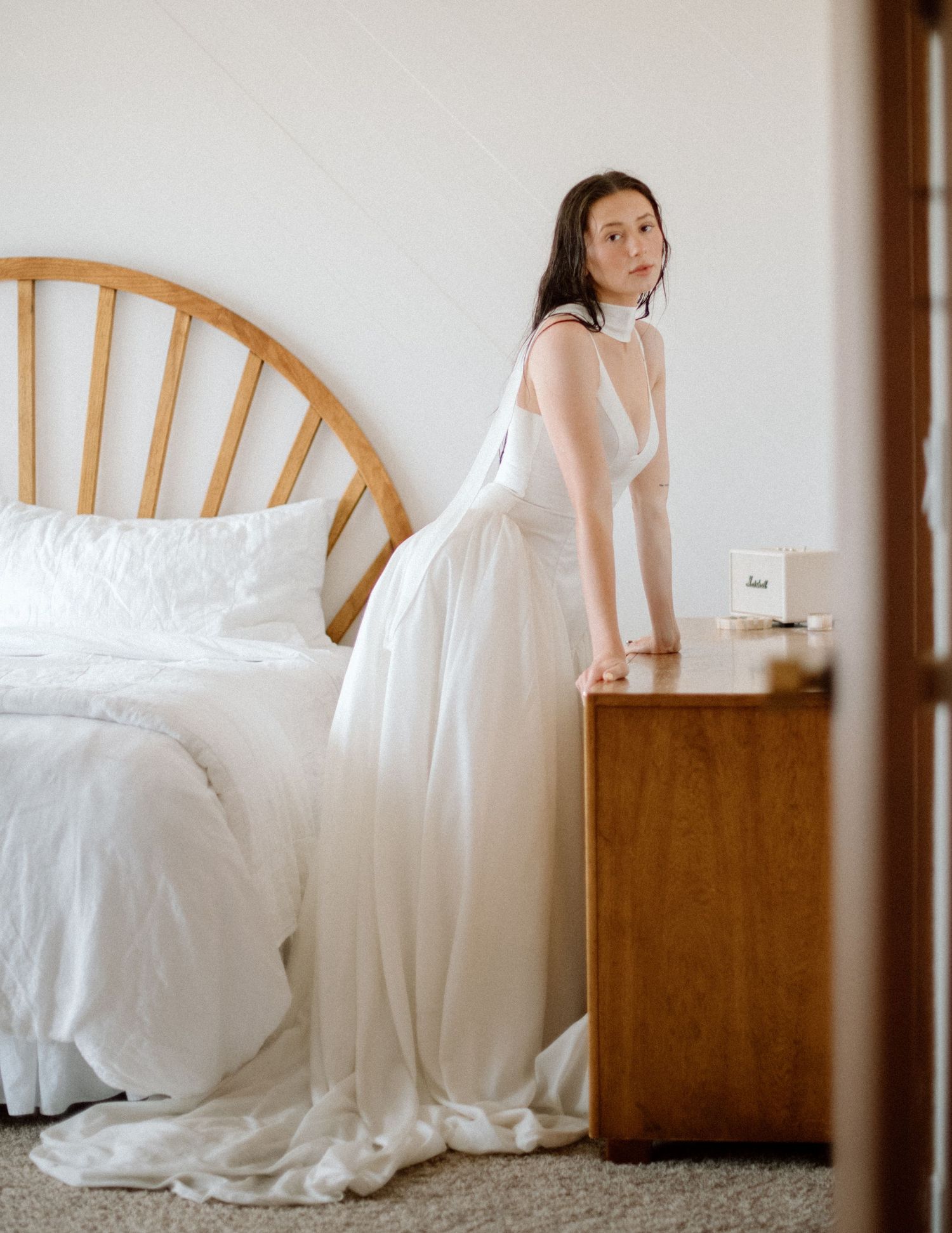 A bridal photo series showing a woman in a white wedding dress seated gracefully against a wooden bed frame.