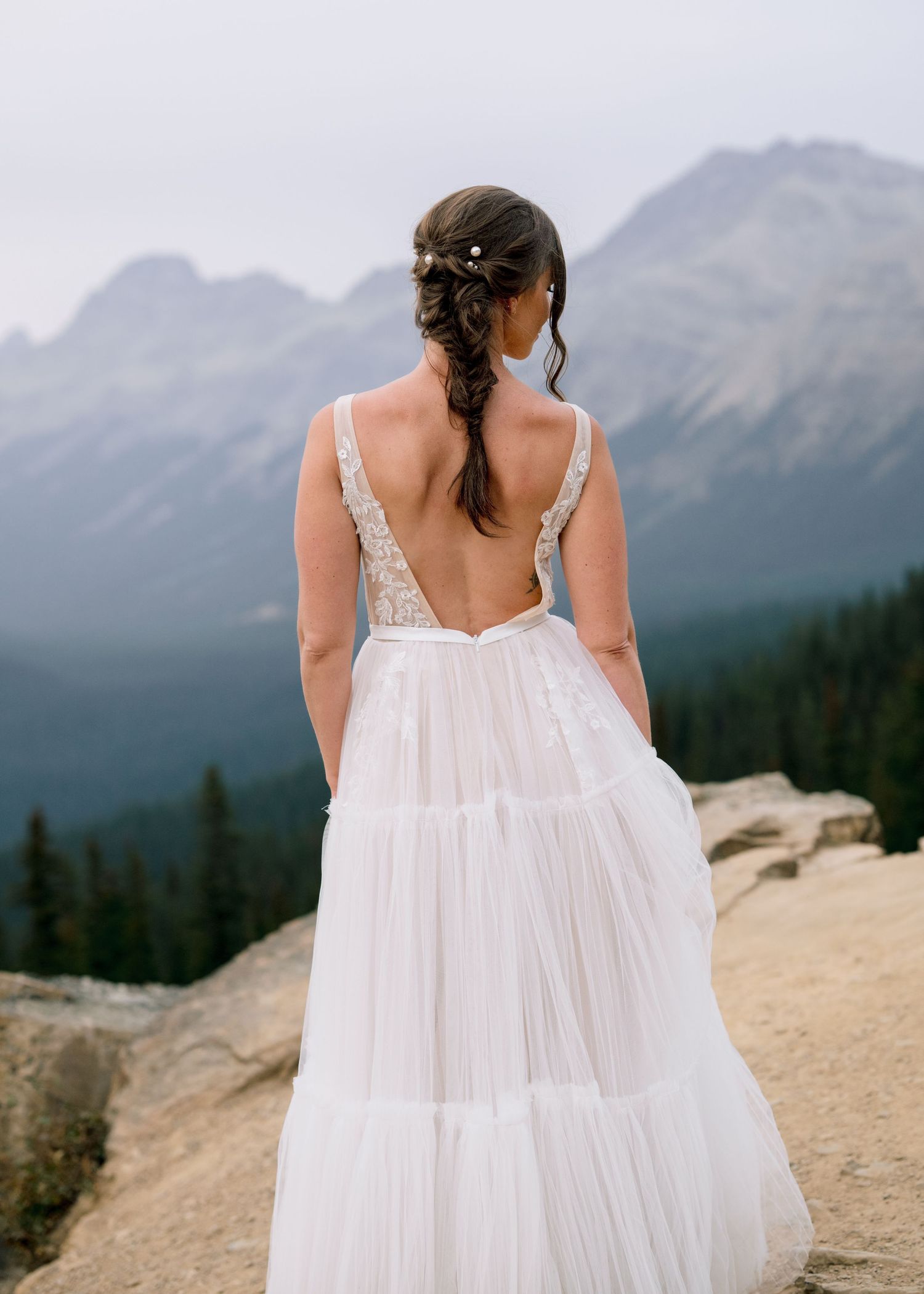 A flowing white wedding dress catches the mountain breeze against a backdrop of majestic peaks and forest.