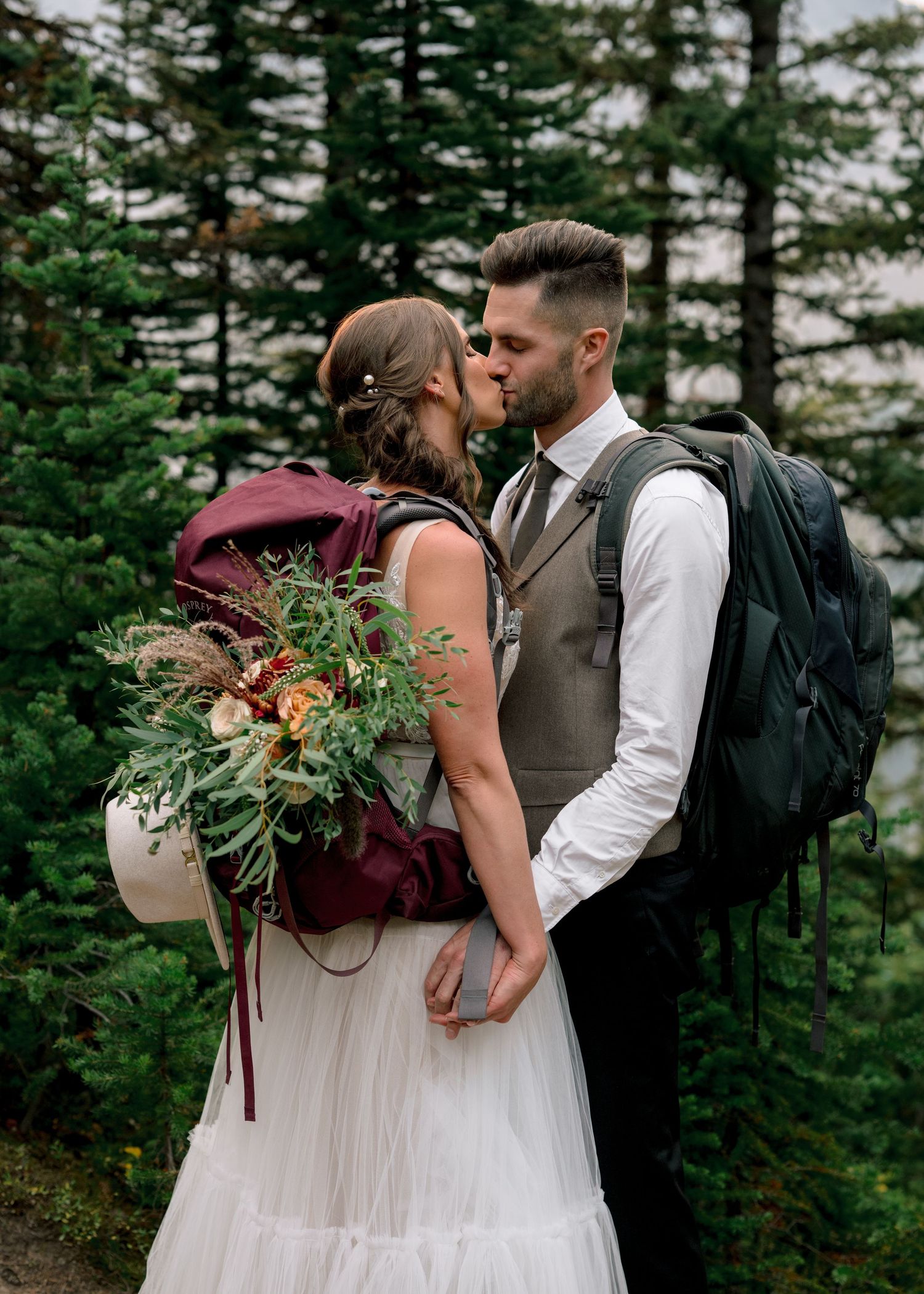 A couple shares an intimate moment while wearing hiking backpacks and formal wear in a forest setting.