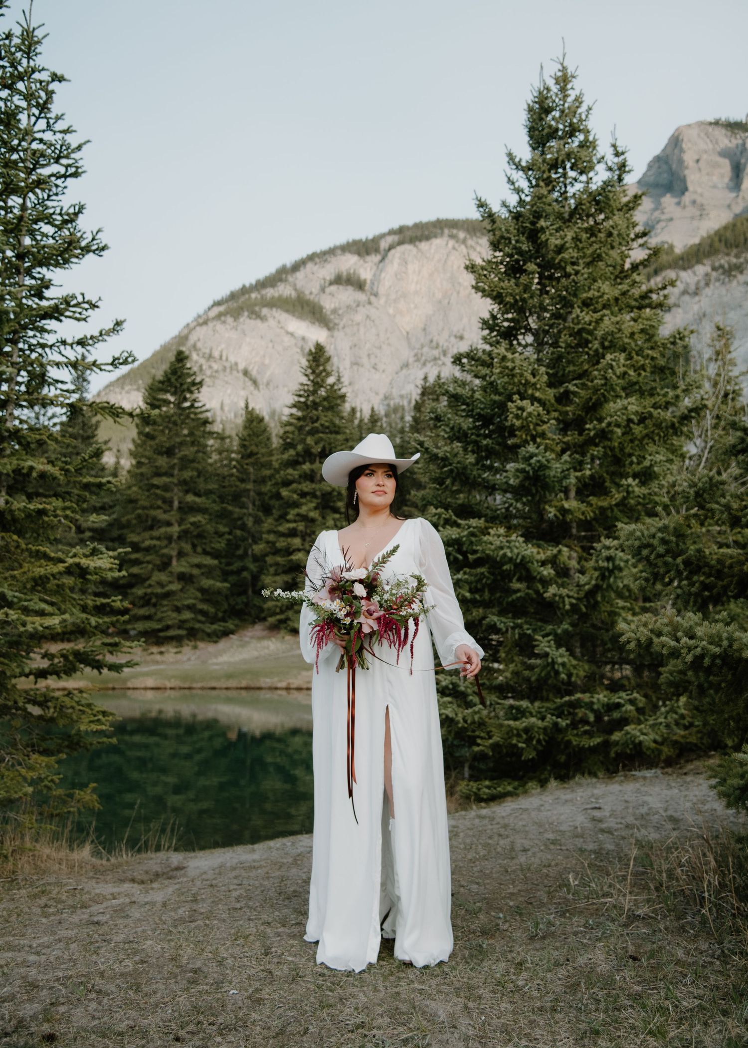 A bride in western attire poses with her bouquet against a dramatic mountain landscape. Banff and Calgary Wedding Photographer