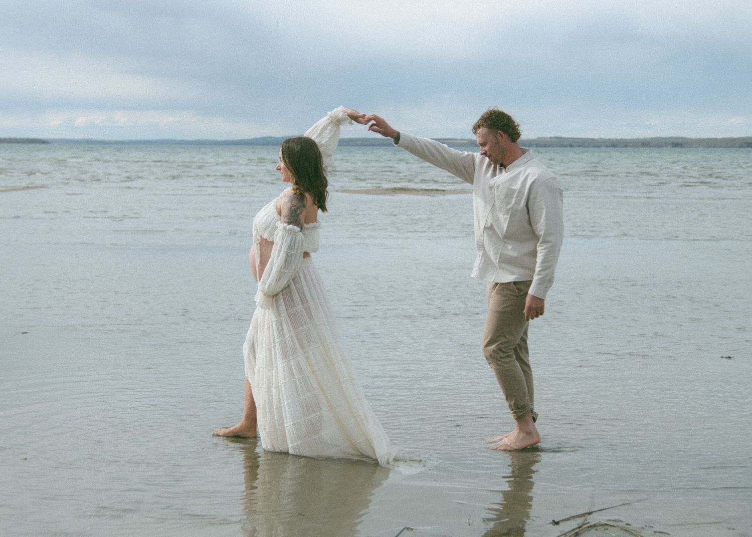 Couple dancing in shallow water wearing white during a beach photoshoot. Calgary Alberta Maternity Photographer.