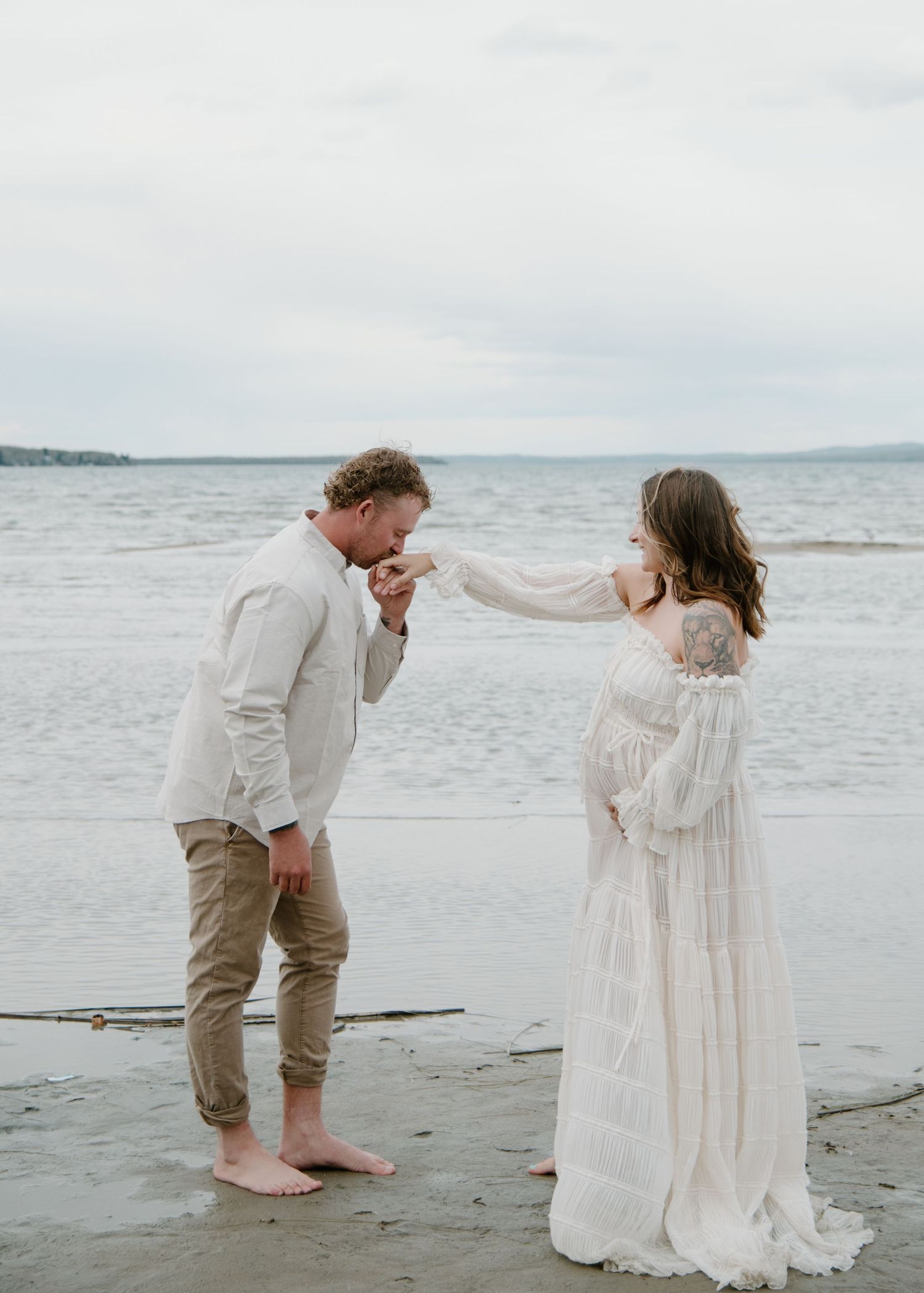 A romantic gesture between a couple wearing light colors on a cloudy beach. Calgary Alberta Maternity Photographer.