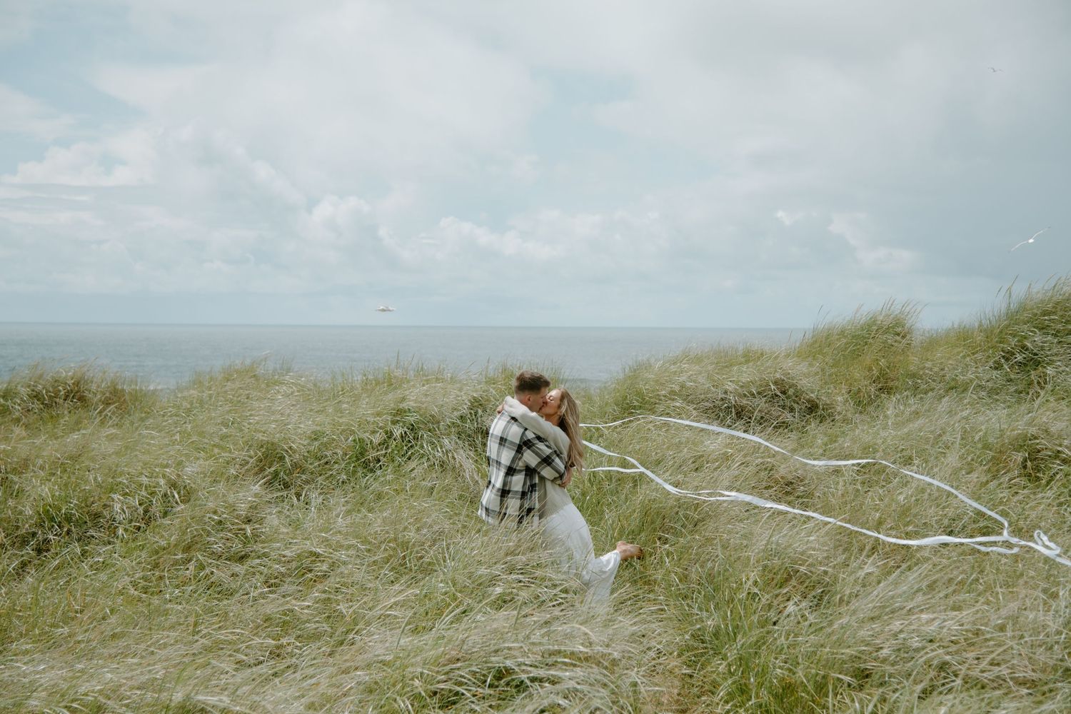 A couple runs through windswept beach grass with flowing white fabric on a cloudy day on Oregon Coast. Calgary Lifestyle and Engagement.