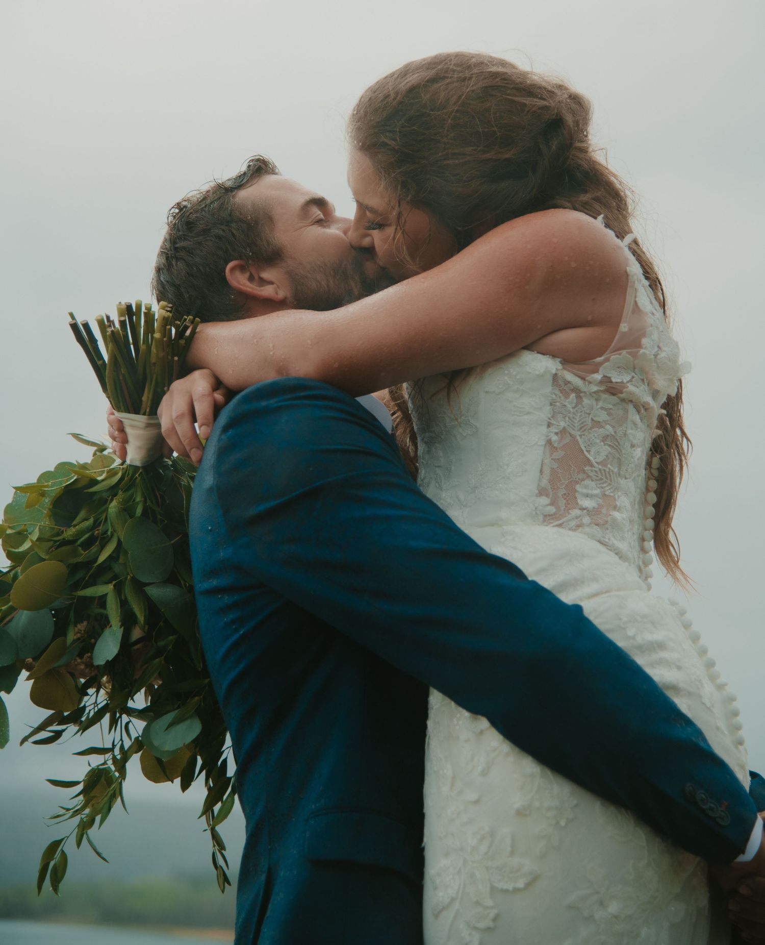 Romantic embrace between newlyweds with bouquet against misty mountain backdrop. Calgary Wedding Photographer
