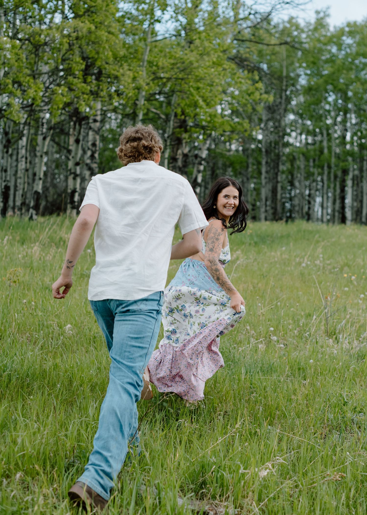 Two people run playfully through a green meadow with aspen trees in the background. Calgary Alberta Engagement Photographer.