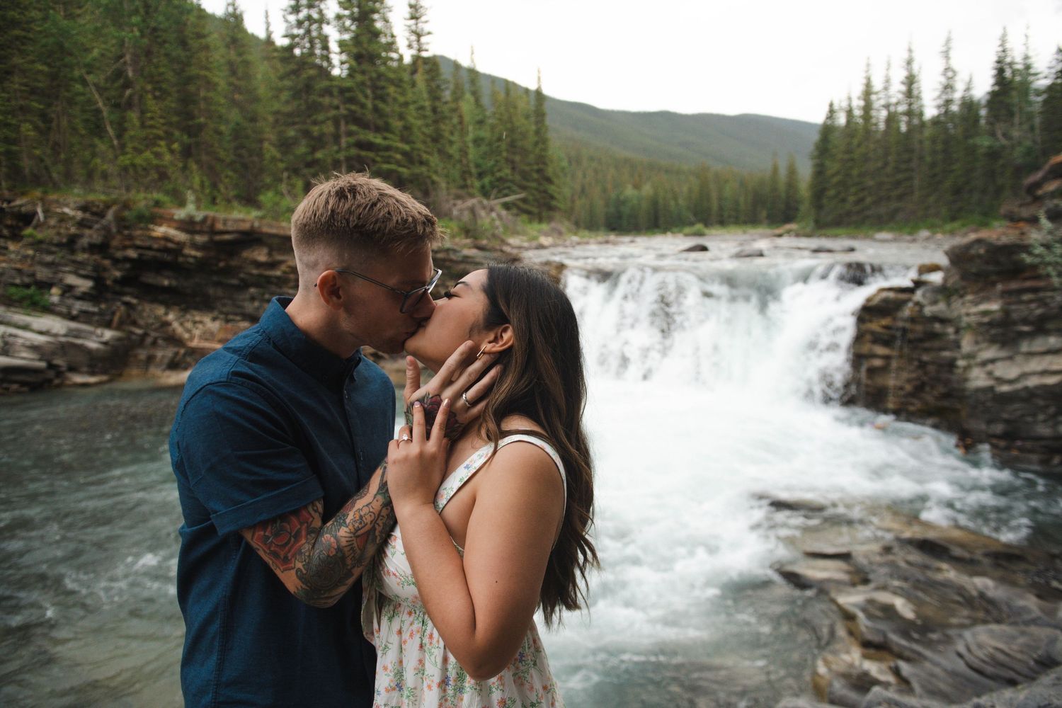 Couple embracing near a scenic waterfall in a forest landscape in Calgary. Calgary Lifestyle and Engagement Photographer.