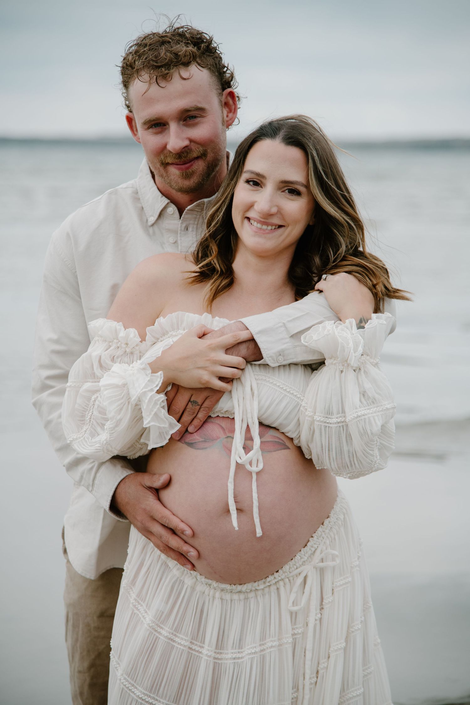 Joyful maternity photography session on overcast beach with couple in light coloured clothing. Calgary Alberta Maternity Photographer.