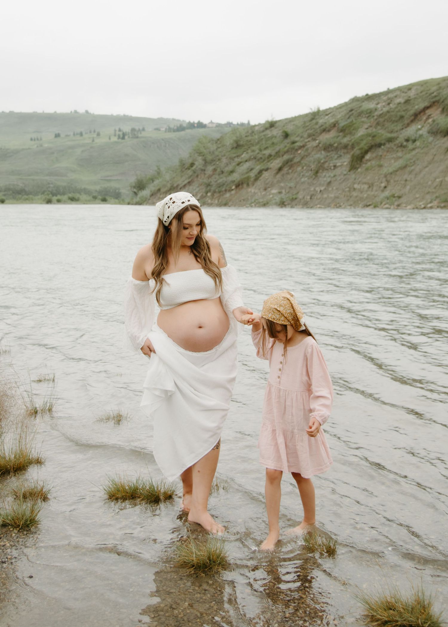 A mother and daughter share a tender moment at the water's edge near rolling hills on an overcast day.