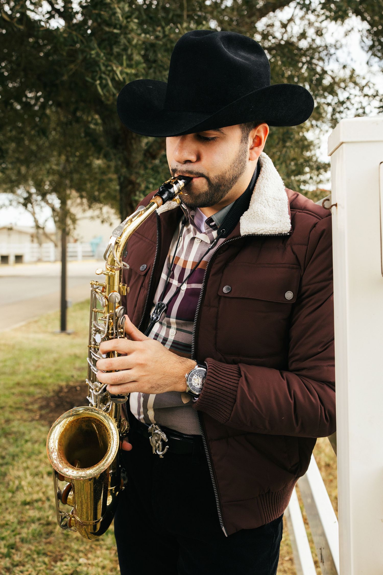 A musician in a brown jacket and cowboy hat plays saxophone outdoors near a white fence.