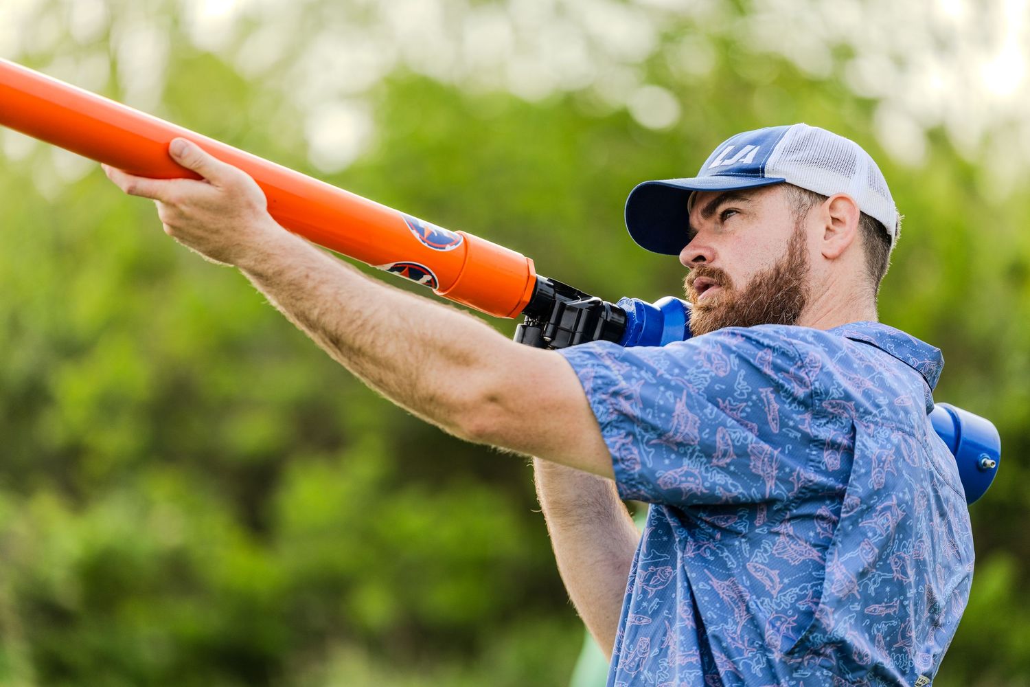 A person wearing a blue tie-dye shirt demonstrates proper form for throwing a clay target with an orange disc.