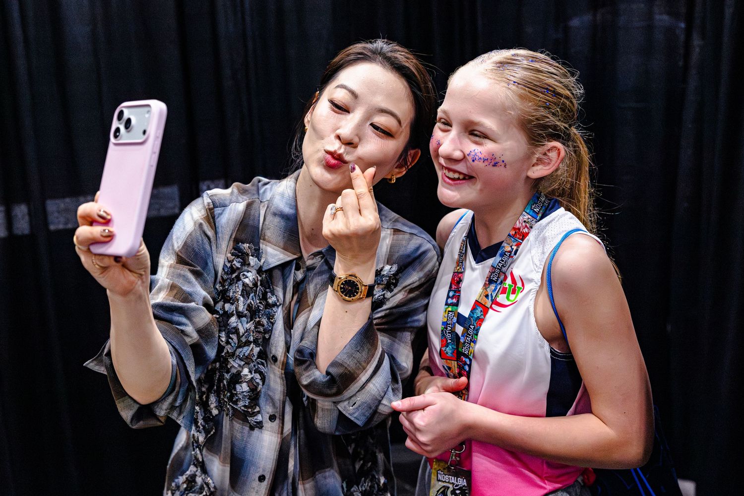 Two women pose for a selfie, one making a heart sign, the other with face paint, at an indoor event.