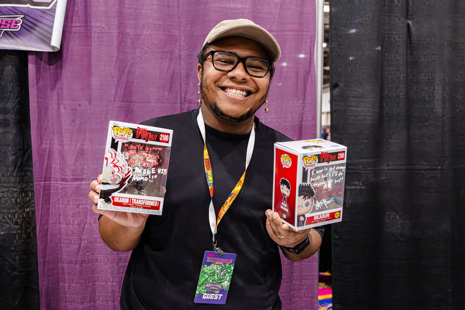 Person smiling at comic con holding two Funko Pop collectible boxes while wearing a lanyard and hat.