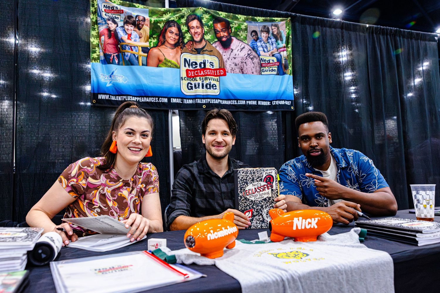 Three smiling people sit at a signing table with Nickelodeon branding at a fan convention event.