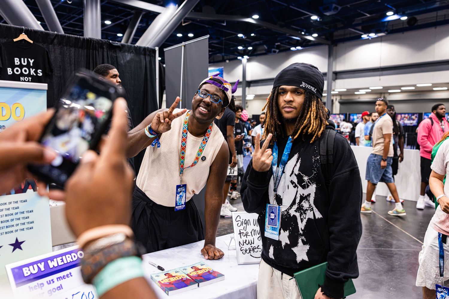 Two young men pose together at a comic convention booth with a Buy Books sign in the background.