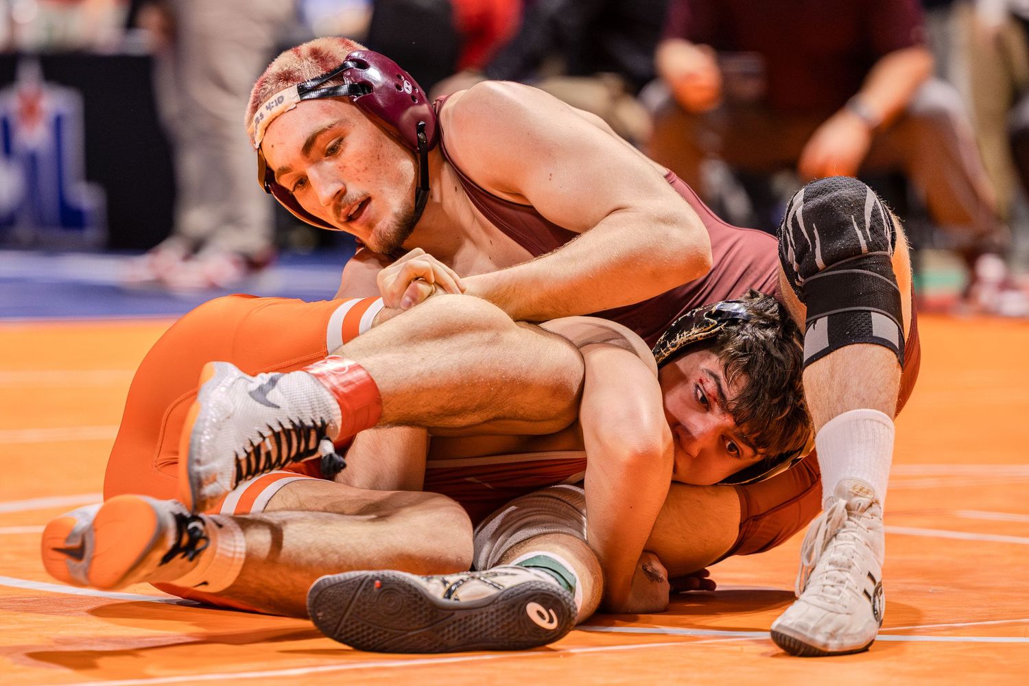 Two wrestlers compete in an intense match on an orange wrestling mat during a competitive bout.