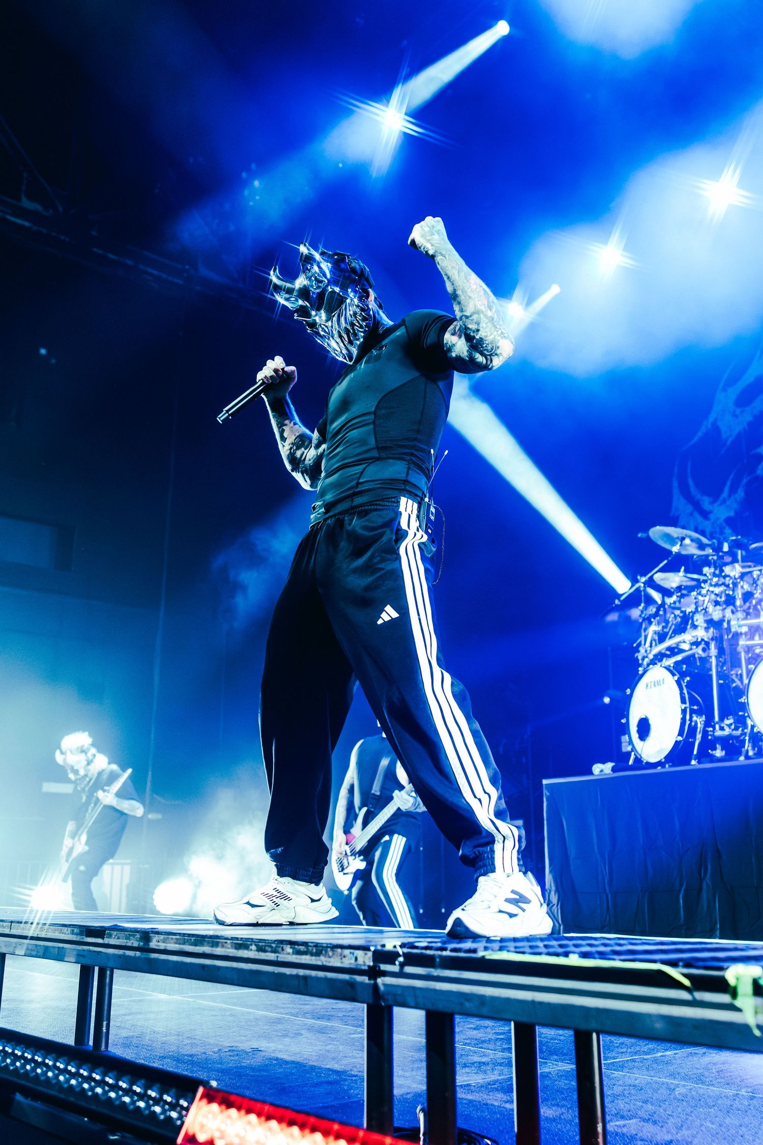 Masked performer in Adidas tracksuit raises fist on stage under dramatic blue lights during live concert.