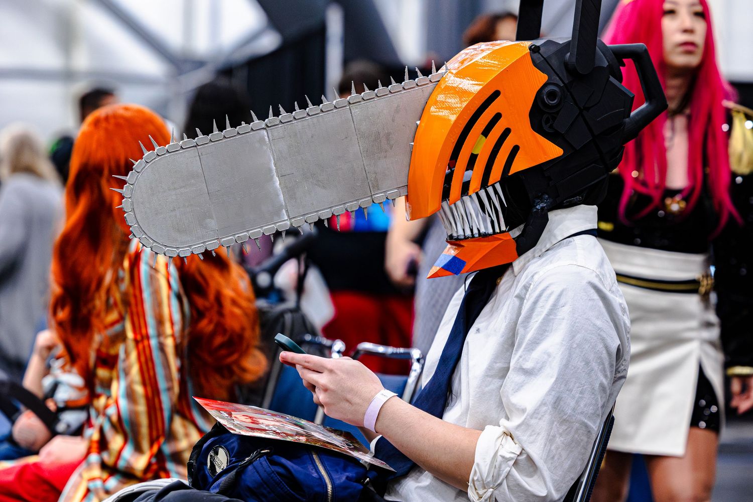 Person wearing orange chainsaw helmet cosplay at anime convention with colorful cosplayers in background.