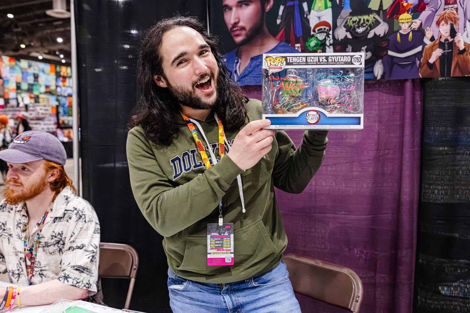 Smiling man with long hair holds Funko Pop box set at a comic convention signing table.