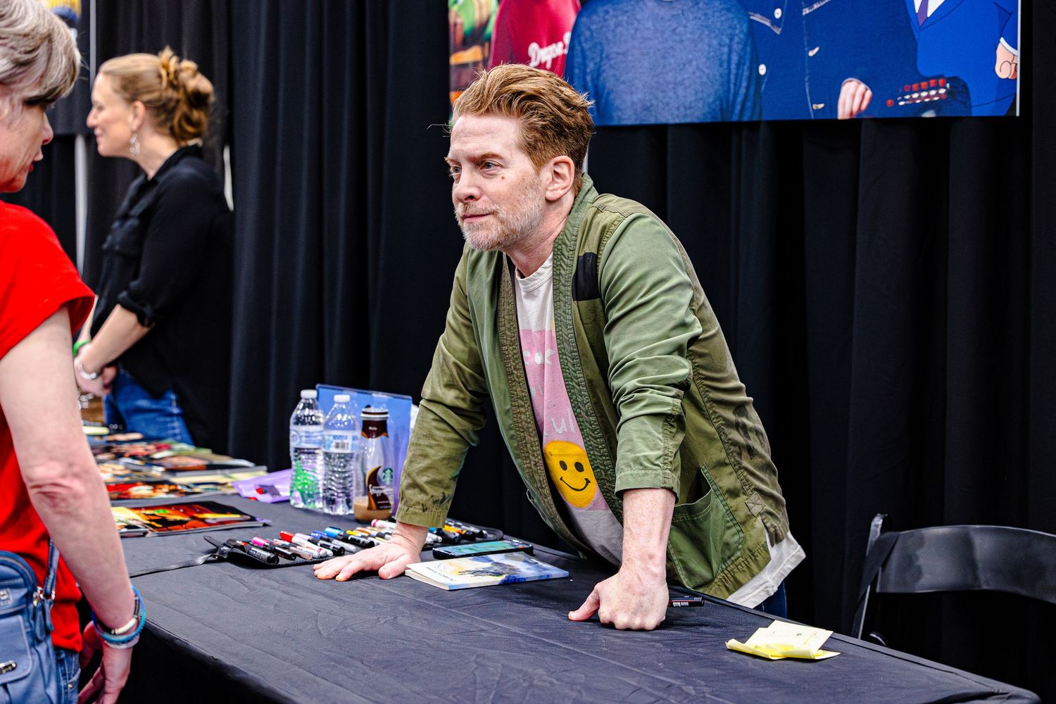 Man in green jacket signing autographs at a convention table with fans waiting in line.