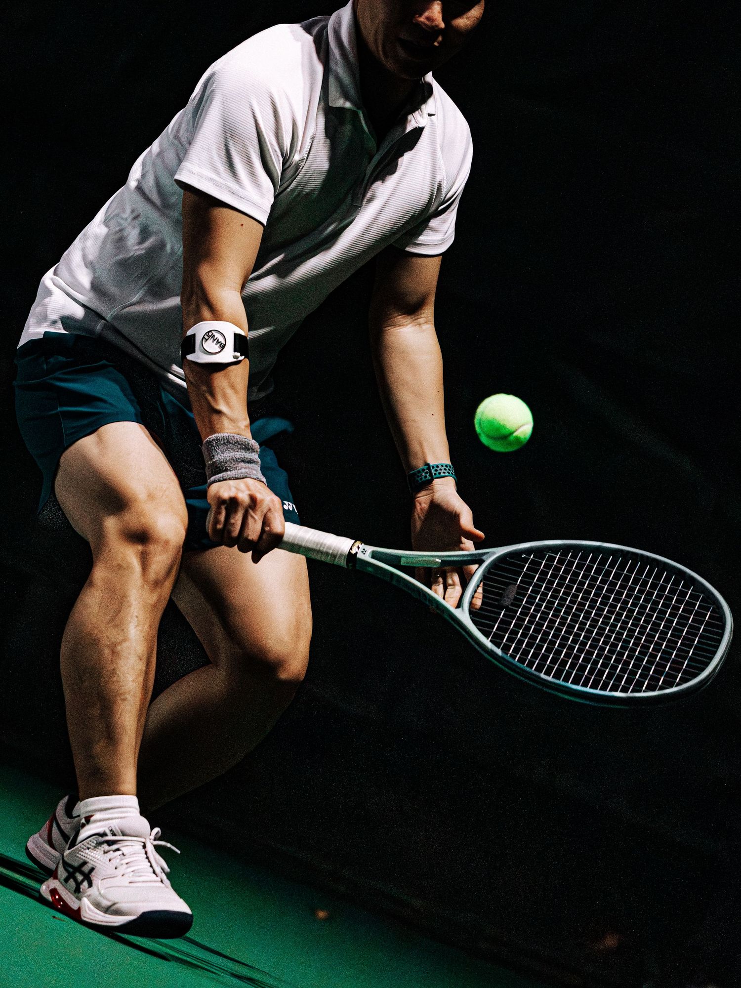 Tennis player in white clothing demonstrates low stance and racquet grip technique on dark court with green surface.