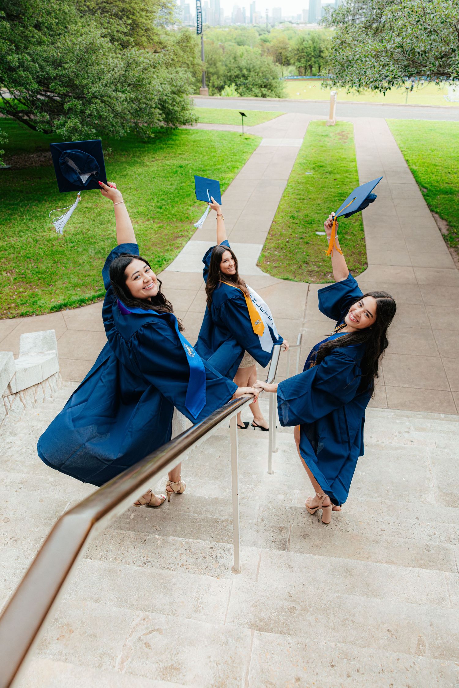 Three women in blue graduation gowns and caps celebrate on outdoor steps, holding caps aloft with green park in background.