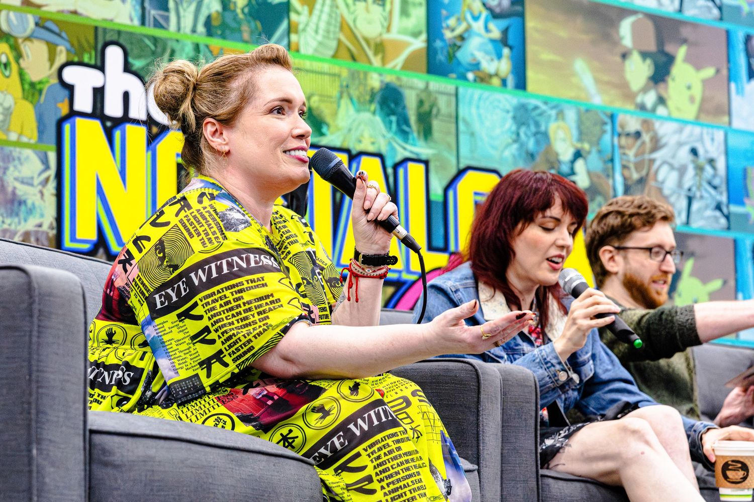 Woman in yellow print dress speaks at comic con panel while others sit beside her on stage.