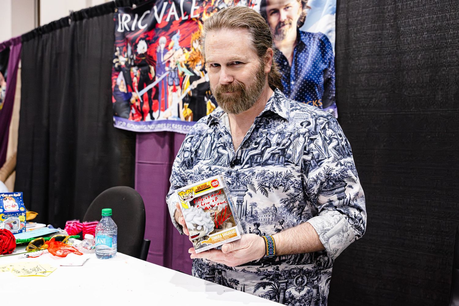 Bearded man in floral shirt holds comic book at Eric Valen signing table at comic convention.